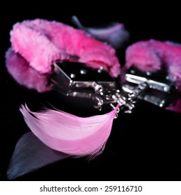 a pair of pink sexy fluffy handcuffs and some pink feathers used as adult toys on a reflecting black background