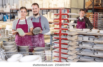 Pair of positive professional artisans, young woman and bearded man in stained burgundy aprons standing in well-lit pottery studio, holding glazed ceramic pieces with pride - Powered by Shutterstock