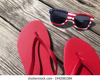 A pair of red flip flops and patriotic sunglasses sitting on a wooden background.