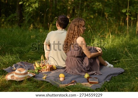 Pair resting back to back beneath trees with picnic and sunset glow