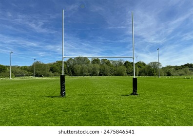 A pair of rugby goal posts stand prominently at the center of a lush, green field under a blue sky. In the background, trees line the edge of the playing area in, Cottingley, UK - Powered by Shutterstock