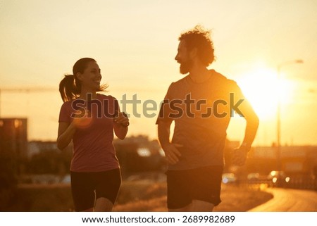 Pair of runners smiles while jogging side by side on a city path at sunset, encouraging fitness and healthy habits in a vibrant outdoor environment.