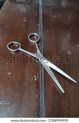 A pair of shiny silver scissors rests open on a dark, worn wooden table surface, ready for various cutting tasks.