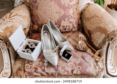 A pair of silver high heels and a box of earrings sit on a pink chair. The chair is covered in a floral pattern - Powered by Shutterstock