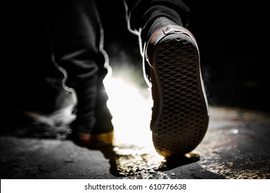 A Pair Of Sneakers Is Seen While A Guy Wearing Jeans Is Walking In The Darkness And A Strong Light Is Illuminating The Darkness In A Close-up Shot. 