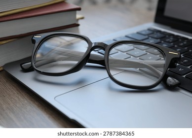 Pair of stylish glasses, laptop and stack of books on wooden table indoors, closeup