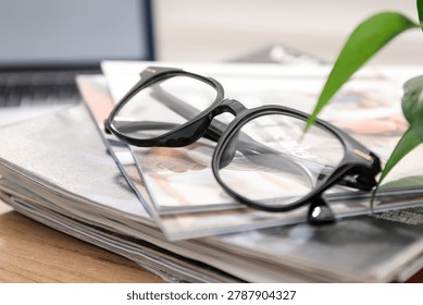 Pair of stylish glasses and stack of magazines on wooden table indoors, closeup