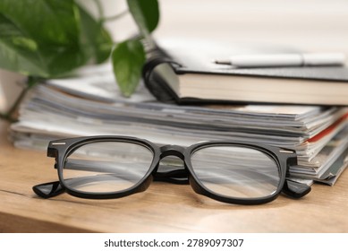Pair of stylish glasses, stack of magazines and notebook on wooden table indoors, closeup
