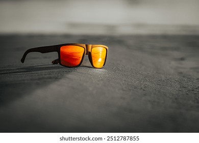 A pair of sunglasses with orange-tinted lenses rests on sandy ground, reflecting a serene beach sunset. The minimal background enhances the focus on the eyewear. - Powered by Shutterstock