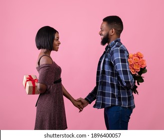 Pair of sweethearts hiding Valentine surprises for each other behind their backs over pink studio background, side view. Passionate black couple with flowers anf gift celebrating lover's holiday