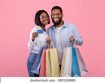 Pair of sweethearts shopping for Valentine's Day, holding gift bags and credit card, promoting contactles payment, pink background. Millennial black couple buying presents on web