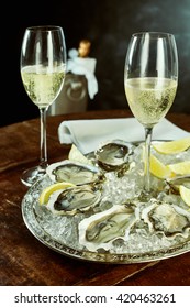 Pair of tall glasses of champagne next to silver platter of oysters and lemon slices on table with cooler and napkin in background
