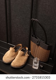 A pair of tan suede clogs sits next to a woven straw tote bag and a glass perfume bottle on a slatted black shelf
