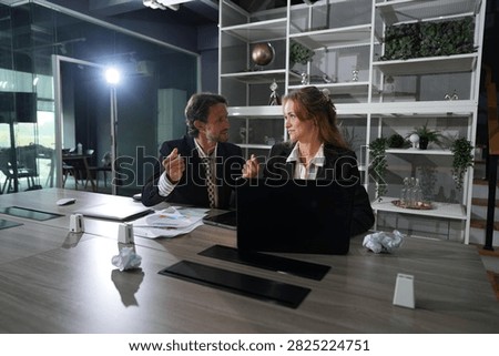 The pair turn from the screen to talk directly, profiles lit by the desk lamp, planning their next move late in the glass walled office in a bright modern coworking office.