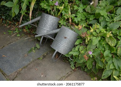 Pair of Two Upside  Down Galvanised Metal Watering Cans on a Flagstone Footpath by the Edge of a Herbaceous Border in a Country Cottage Garden in Rural Devon, England, UK