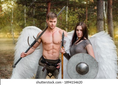 A pair of warrior angels, an athletic man and woman in armor with weapons in their hands and white wings behind their backs against the backdrop of the park.