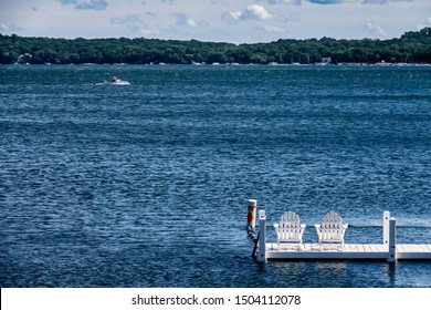 Pair of white Adirondack chairs at end of narrow white dock on large, rippled lake where a small motorboat is passing by on a breezy day in summer, with light digital oil-painting effect