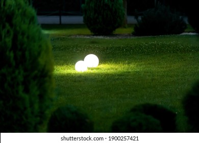 A Pair Of White Ball-shaped Lighting Fixtures On A Green Lawn Illuminate The Landscape Of A Spring Evening Park With Thuja Bushes, The Glow Of Ground Lanterns, Nobody.