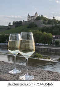 Pair Of White Wine Glasses On Stone Wall With Landscape In The Background Of A Wurzburg Wine Estate And Its Vineyards In The Northern Bavaria Region, Germany.