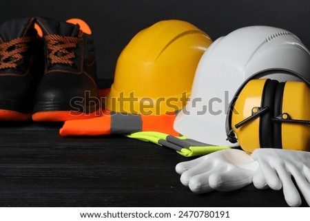 Pair of working boots, hard hats, protective gloves, earmuffs and reflective vests on black wooden surface, closeup