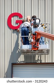 A Pair Of Workmen Install A Sign On The Side Of An Industrial Building Using A Lift.