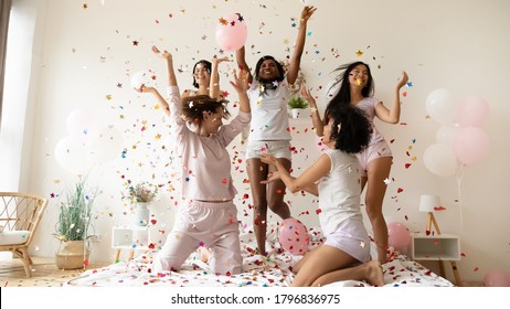 Pajama or bachelorette party celebration concept. Five slim active multiethnic women wearing comfy nightwear sexy glamor pyjamas hanging out jumping dancing on bed under falling multi colored confetti