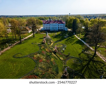 Pakruojis Manor stands with its white facade and red roof, surrounded by lush gardens. Vibrant tulip beds and tall trees enhance the expansive green lawn. - Powered by Shutterstock