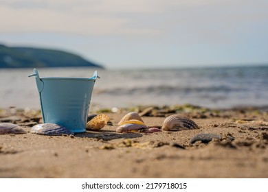 Pale Blue Bucket Of Collected Sea Shells On A Sandy Beach