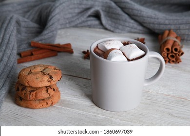 Pale mug of chocolate with marshmallow, spices, cookies, grey knitted scarf on white wooden table background. Winter drink concept. Spicy cocoa, menu, closeup, copy space