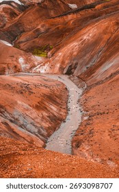 A pale stream winds through rust red rhyolite hills in Iceland, with mineral streaks, green moss patches, and warm late afternoon light accenting orange and ochre tones.