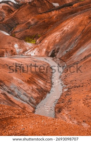 A pale stream winds through rust red rhyolite hills in Iceland, with mineral streaks, green moss patches, and warm late afternoon light accenting orange and ochre tones.