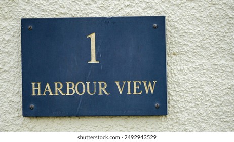 A pale yellow stone wall with a rectangular slate plaque with number 1 harbour view etched on it. One of the many coastal and beach inspired house names in the seaside resort of Tenby, Wales. 