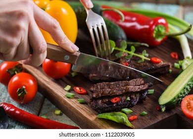 
Paleo Diet. Woman Hands Cutting Well Done Steak On Oak Board Served With Vegetables Close Up