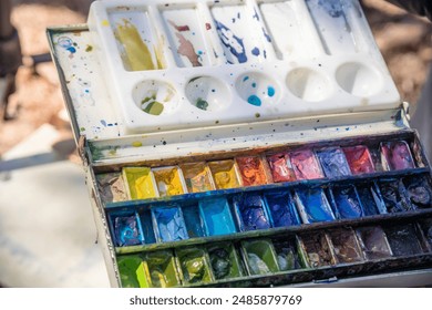A palette of watercolors with a brush on top. The palette is full of different colors and the brush is ready to be used. Scene is creative and artistic