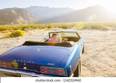 Palm Springs, CA / USA - 05/17/2017: Man And Woman Couple Driving Off Into Sunset In Blue Convertible In The Desert At Sunset