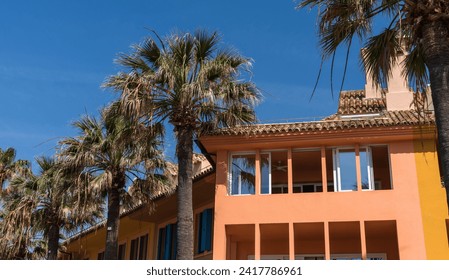Palm trees sway before a colorful, sunlit Mediterranean building with terracotta tiles, evoking a warm, tropical atmosphere. - Powered by Shutterstock