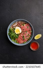 Pan Asian Style Soup On A Spicy Sturdy Beef Broth With Boiled Eggs, Red Onion, Cilantro And Thin Slices Of Marbled Beef Over Black Concrete Background. Overhead View