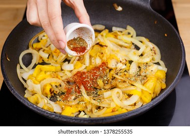 Pan cooking on a ceramic hob on a wooden background kitchen table with jackfruit and onion inside while a hand pours a small bowl full of powder spices like curry, cumin, oregano or nutmeg.