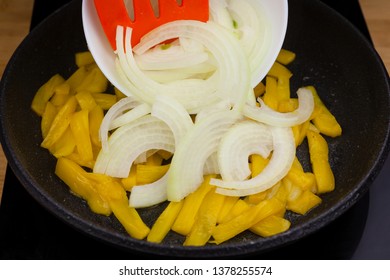 Pan cooking on a ceramic hob on a wooden background kitchen table with chopped onion inside while a hand pours a bowl full of onion.
