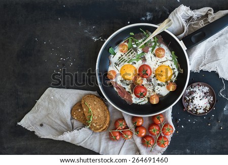 Pan of fried eggs, bacon and cherry-tomatoes with bread on dark table surface, top view