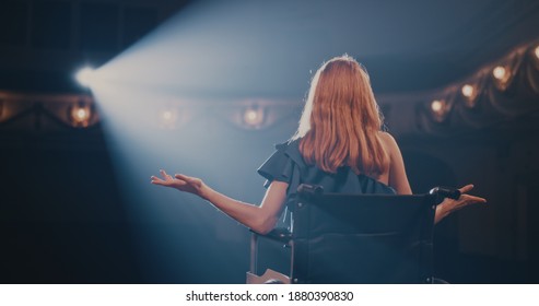 Pan left view of young woman smiling and gesticulating while sitting on wheelchair in spotlight and talking to audience during performance in dark theater - Shutterstock ID 1880390830
