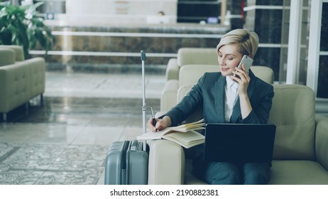 Pan shot of blonde busy businesswoman sitting in armchair in hotel lobby talking mobile phone, writing in notepad and using laptop computer smiling. Business, travel and people concept - Shutterstock ID 2190882381
