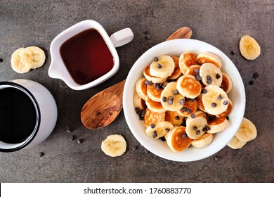 Pancake cereal with bananas and chocolate chips. Mini pancakes in a bowl, top view table scene over a dark background. Trendy breakfast food concept.