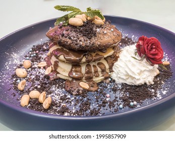 Pancakes Covered In Chocolate Hazelnut Sauce, Crushed Oreos, Peanuts And Whipped Cream. Stack Of Pancakes On Black Plate. Unhealthy Breakfast Served In Restaurant With Powder Sugar And Rose Decoration