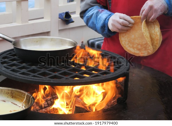 Pancakes from the dough are fried in a pan over an open\
fire. A few pancake pans stand on the barbecue grill over coals\
with flames. Pancakes are baked at an outdoor party.               \
         