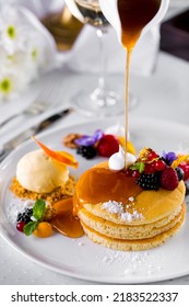 Pancakes With Maple Syrup From A Ceramic Gravy Boat On A White Plate With Edewiki Berries, Raspberries, Red Currants, Cookie Crumbs, And Vanilla Ice Cream. A White, Ceramic Plate Stands On A White Table