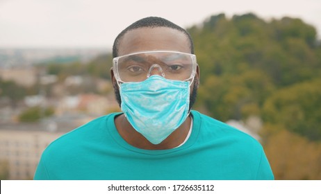 Pandemic 2020. Portrait Of African Healthy Man Wearing Protective Face Mask And Goggles Looking Straight Standing On The Hilltop Above City. Quarantine. Safety Care.