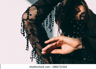 Pandemic Fashion. Night Club Party. Gypsy Style. Woman In Black Sheer Dress Decorated Crystal Beads Face Mask Looking At Camera On White Shadow Light Wall.