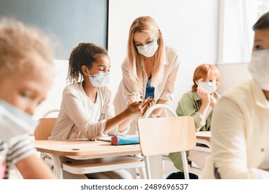 Pandemic. Sanitary measures against Covid19 coronavirus spread at school. Teacher using sanitizer with her students pupils schoolchildren before lesson class wearing protective face masks. - Powered by Shutterstock