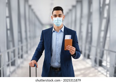 Pandemic Travels. Portrait Of Young Arab Man Wearing Protective Medical Mask Standing In Airport Terminal, Male Traveller Holding Passport And Flight Tickets, Middle Eastern Guy Having Business Trip - Powered by Shutterstock
